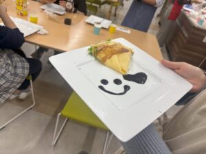 両手で四角い白いお皿を持っているところ。お皿のはしの方に、扇形に畳まれたクレープが盛り付けられ、お皿中央にチョコレートでスマイルとハートのイラストが描かれている。