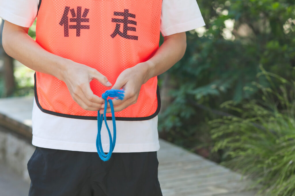 「伴走」と書かれたオレンジ色のビブスを着た人の上半身のクローズアップ。伴走用のたすきを手に持っている。たすきはダランと垂れ下がっている。