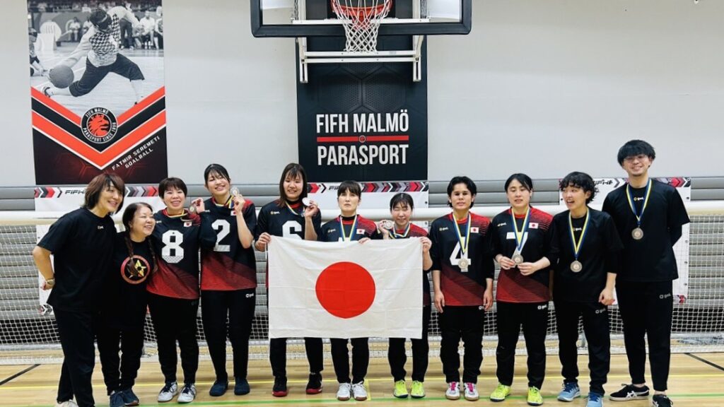 横一列に並んで立つオリオンJAPAN女子チームの選手7人。銀メダルを首からかけていたり、手に持ったりしている。その両隣には4人のコーチやトレーナーが立っている。真ん中の3人は大きな日の丸の旗を持っている。