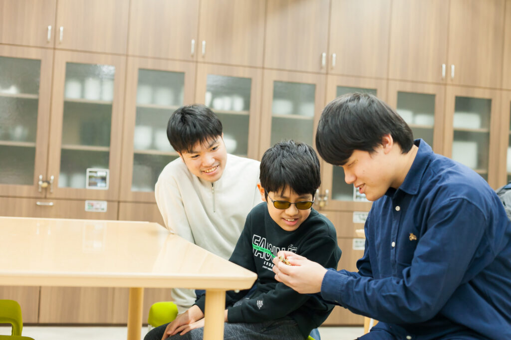 川本さんが何か手に持って指先で触っている様子を、視覚障害の小学生の男の子とガイドヘルパーが横から覗き込んで笑顔で話している。