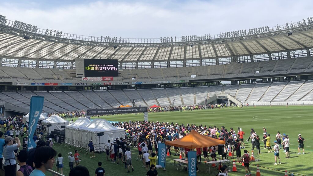 参加したリレーマラソンのスタジアムの様子。たくさんの参加者がトラックに集合している。「味の素スタジアム」と書かれた大型ビジョンがあり、青空も見えている。