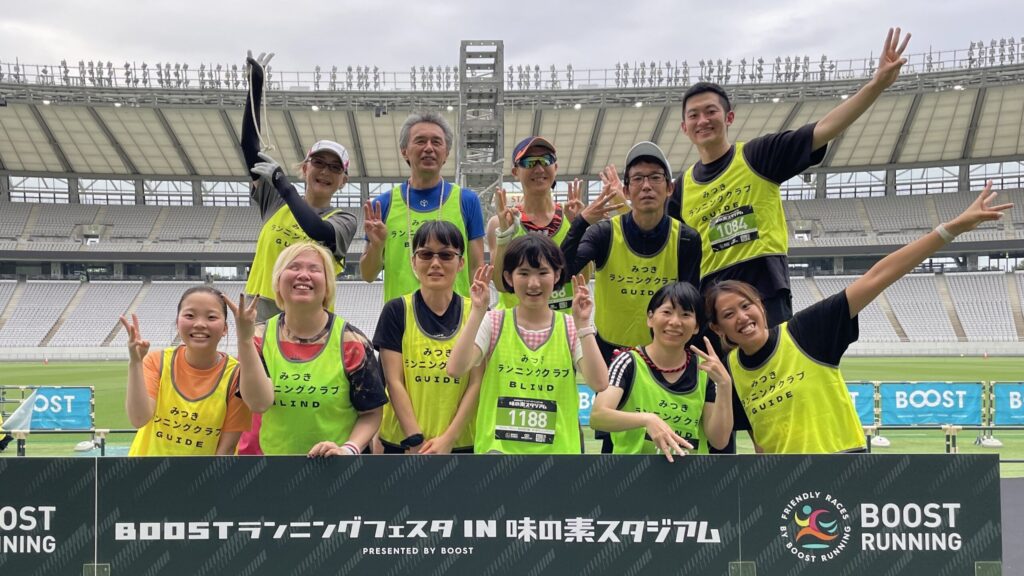 大会参加時の集合写真。「BOOSTランニングフェスタIN味の素スタジアム」と書かれたパネルの後ろに立って、笑顔でポーズをしているメンバーの皆さん。