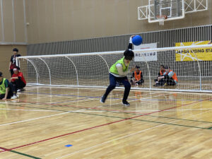 ゴールボール競技中の様子。ゴール前でボールを投げようと、前かがみで右腕を大きく後ろに引いている選手と、守りの準備をしている二人の選手が見える。ゴール裏やゴールわきに審判やほかの選手もいる。