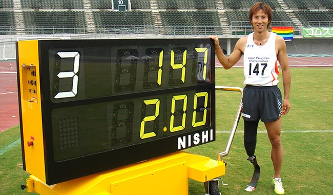 競技場でレースクロックに右手をかけ、に笑顔で立っている鈴木さん。右足は競技用の義足。