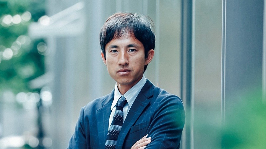 屋外で、ジャケットとネクタイ姿で腕を組んでいる鈴木さん。