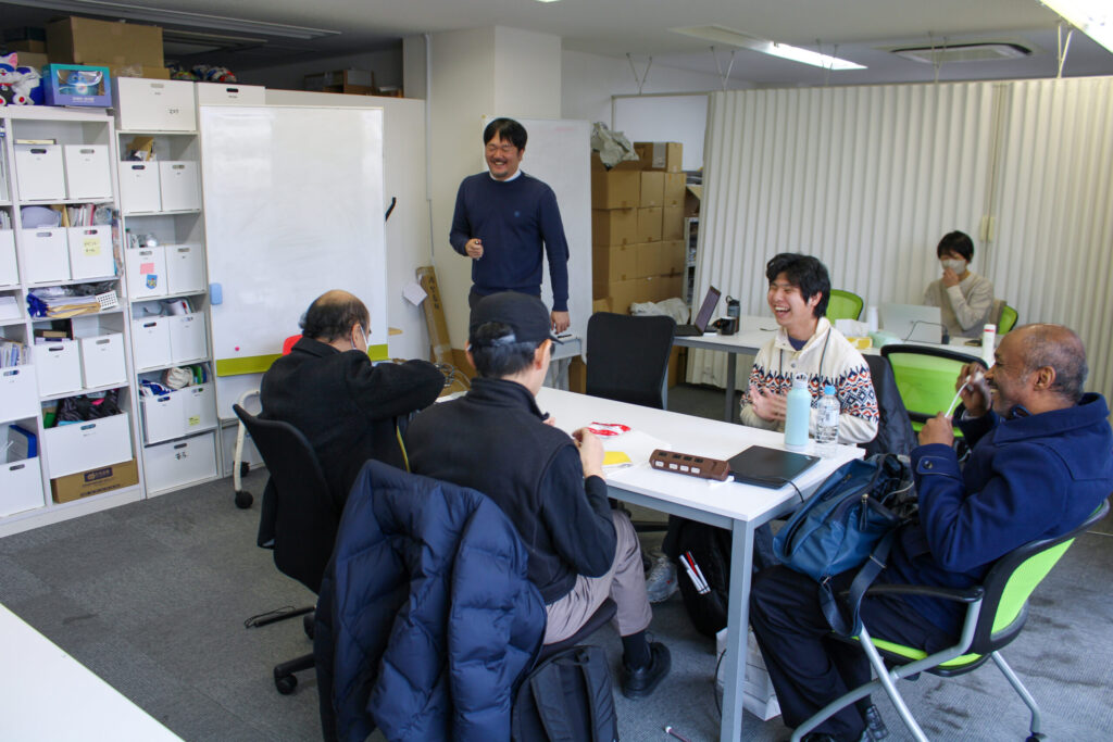 会議室で、机を囲んで笑顔で話し合っている5人の男性。4人は座っていて、ファシリテーターの大坪さんは、ホワイトボードの前に立っている。