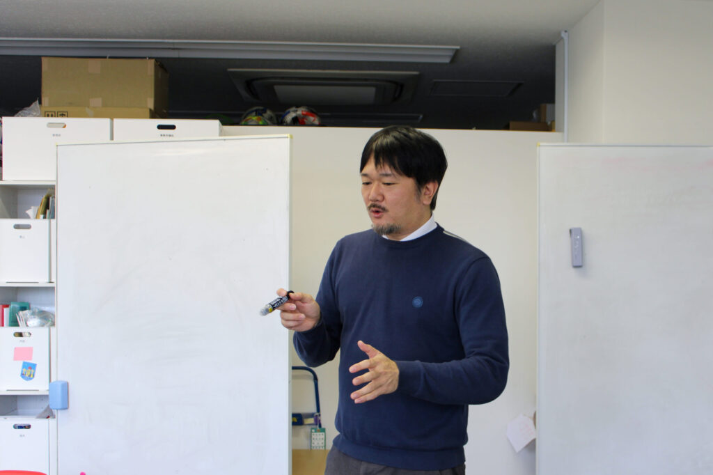 紺色のサマーセーターを着た大坪さんが、手ぶりを交えて真剣に話す様子。右手にホワイトボード用のペンを持っている。