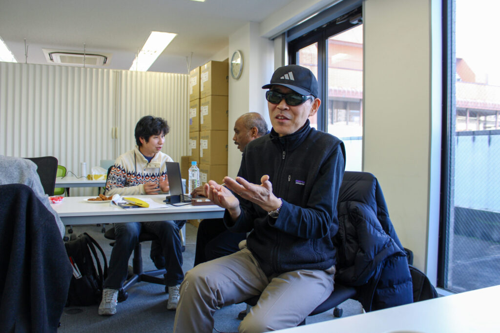 椅子に座っている参加者が、笑顔で手ぶりを交えてこちらを向いて話している。背景には別の参加者二人が真剣に話し合っている様子も見える。