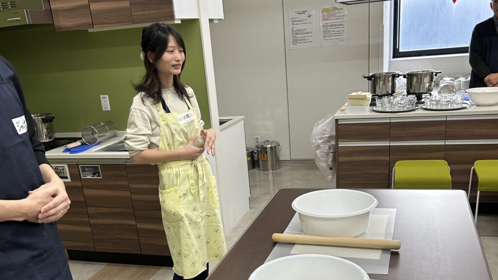 開始前に説明をしているところ。黄色いエプロン姿の「みーちゃん」が、全体に向かって笑顔で話している。