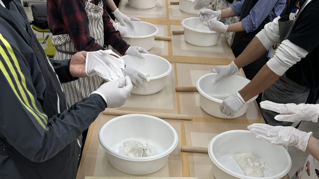 そば粉をこねている様子。テーブルの両側に8人以上が向かい合っている。各自のボウルの中の生地は団子状にまとまっている。