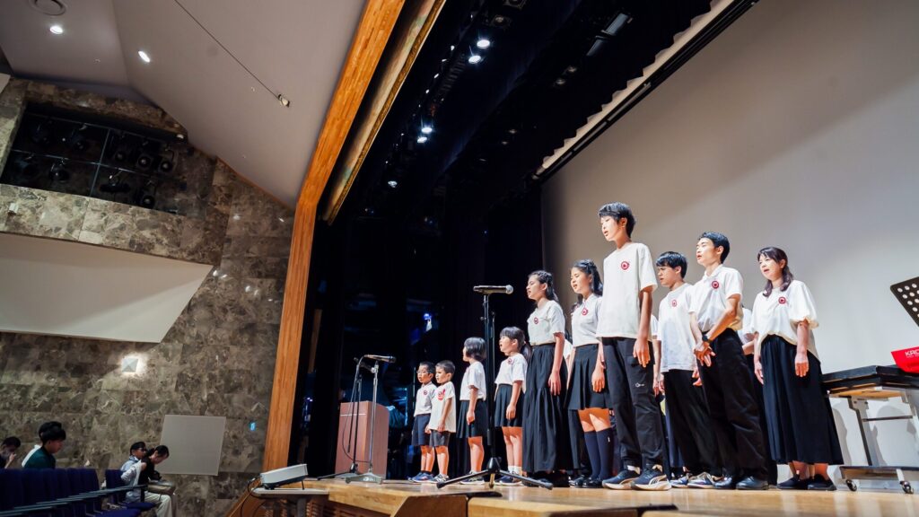 ステージ上で歌ういちご合唱団の皆さんをステージ左下から見上げるように撮った写真。ステージ上の照明が見える。