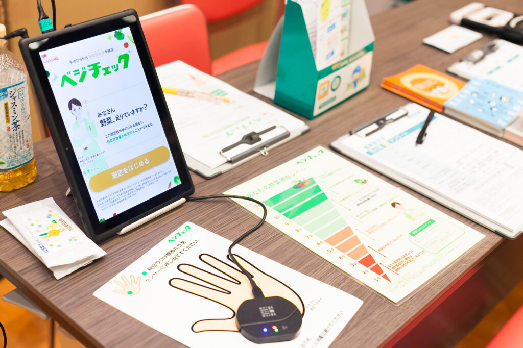 「ベジチェック」と書かれた画面が表示されているタブレットや、手を置いて健康状態をチェックできるデジタル機器、質問票などが置かれた健康チェックコーナーの机の様子。