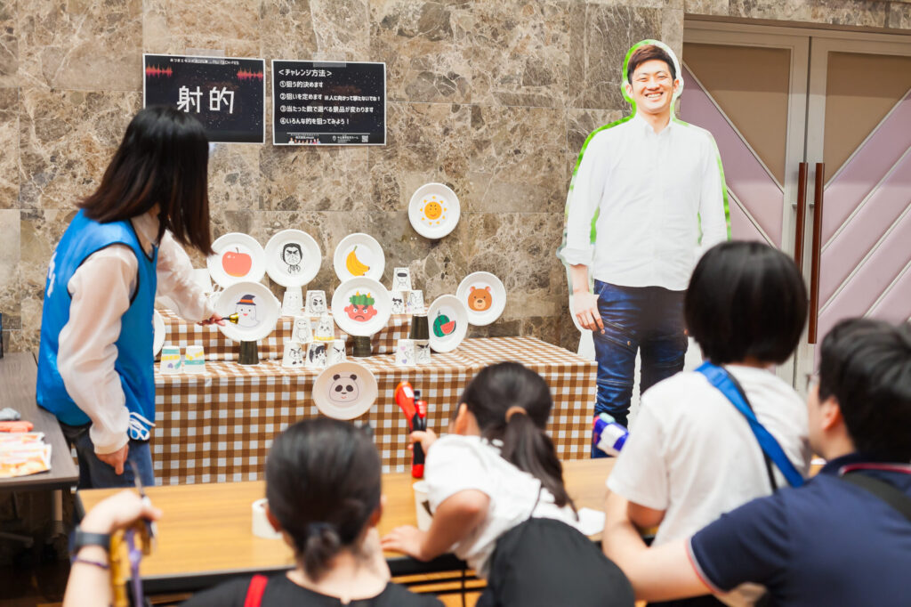 射的コーナーの様子。青いビブスを着たスタッフがマトの前で鈴を振っている。小学生くらいの女の子が、身を乗り出してマトに向かって射的の狙いを定めている。