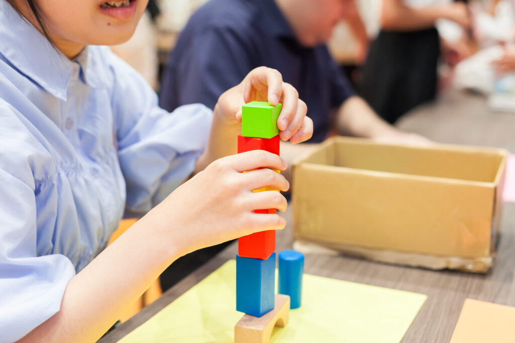 机の上で、ミニサイズの積み木を両手で積んでいる様子。6段以上は積みあがっている。