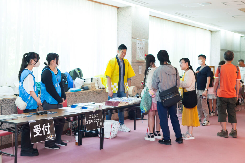 受付横のガラガラ抽選会の様子。黄色いハッピの高橋さんが、参加者にご案内をしている。机には景品がたくさん置かれている。