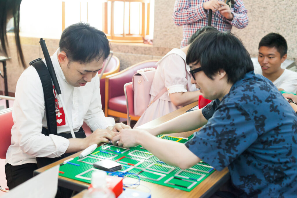 白い服を着て、白杖を手元に持っている男性が、腰かけてカードゲームを体験しているところ。担当者が手を添えてガイドをしんがら説明をしている。