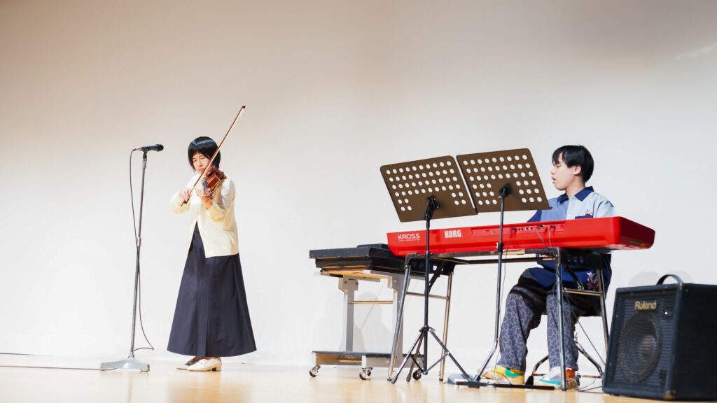 ステージ上でバイオリンを弾いている女性と、その左側で電子ピアノを演奏している男性。