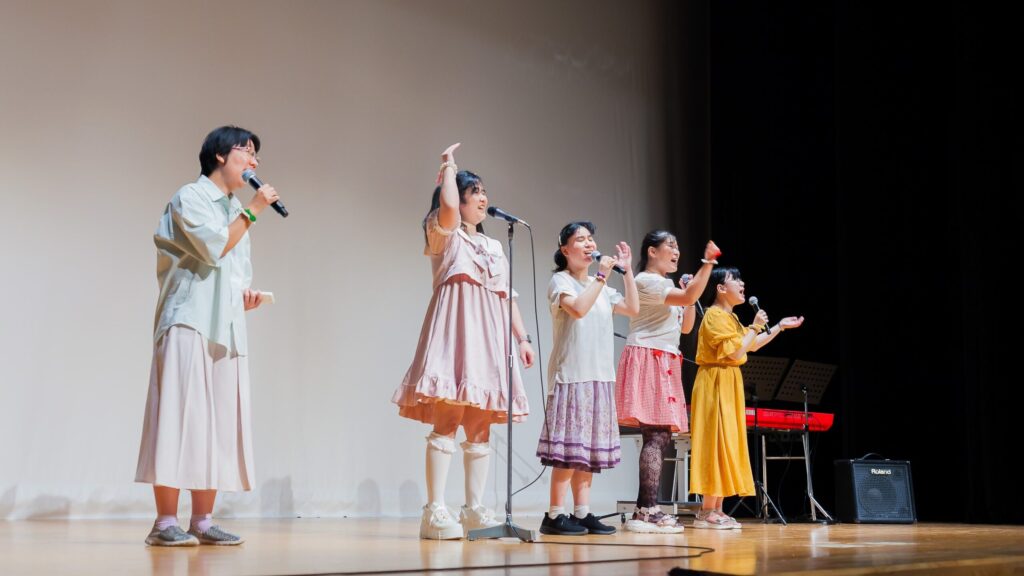 それぞれ違った色合いの衣装を着た女性5人組がステージで歌っている。それぞれ、黄色、赤、紫、ピンク、ミントグリーンの服を着ている。