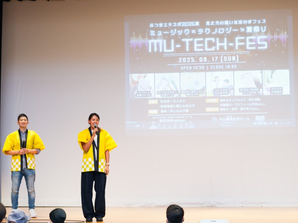 舞台の上でマイクを持って話しているみつき代表の高橋さんと運営の浜さん。背景には「ミューテックフェス」のタイトルが書かれたスライドが投影されている。