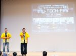 舞台の上でマイクを持って話しているみつき代表の高橋さんと運営の浜さん。背景には「ミューテックフェス」のタイトルが書かれたスライドが投影されている。