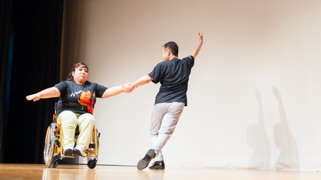 ステージ上で車いすダンスを披露する男女二人組。車いすに乗った女性の左手を左手で握った男性が右手を大きく振り上げてポーズしている。車いすの女性も右手を広げてポーズをとっている。