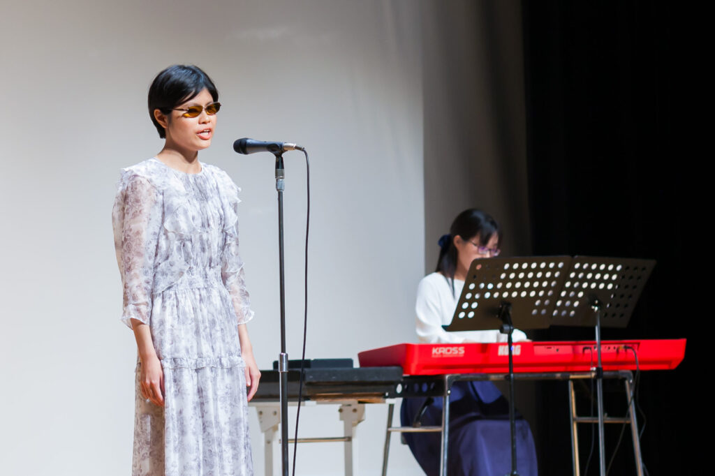 ステージ上で歌う白いワンピースを着た小汐さんと、赤い電子ピアノで伴奏をしているご友人。