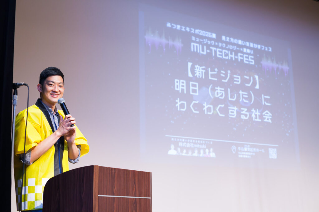 ステージで、マイクを手に新ビジョンを発表する高橋さん。ステージ背景には、新ビジョンの「明日にわくわくする社会」が大きく表示されている。