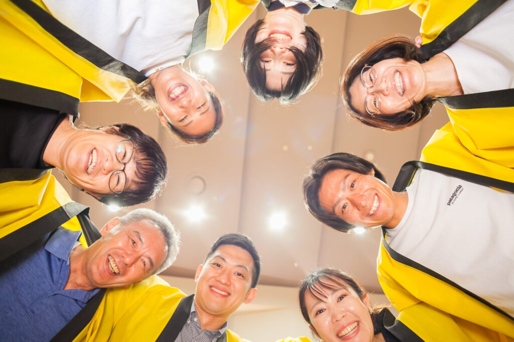 みつきの主要運営メンバー8人が、黄色いハッピを着て円陣を組んでいる様子を真下からとらえた写真。皆さん明るい笑顔。