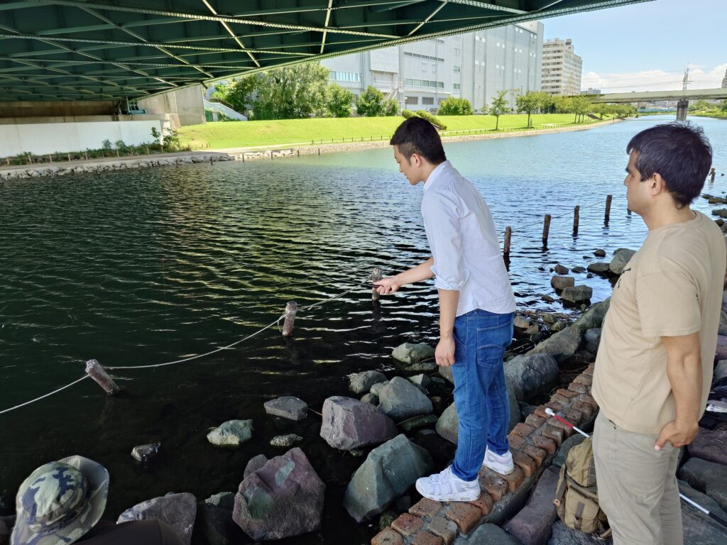 川に向かい釣り糸を垂らして待っているSpotlite代表の高橋さんと、後ろから見守っている筆者ケビンさん。