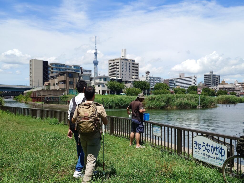 良く晴れている昼間の川のほとり。「きゅうなかがわ」とひらがなで書かれた看板がある柵のよこで、川の様子を確認している男性。その後ろには白杖を持った筆者とSpotlite代表の高橋さん。川の向こう側には東京スカイツリーも見えている。