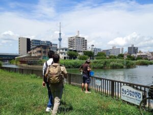 良く晴れている昼間の川のほとり。「きゅうなかがわ」とひらがなで書かれた看板がある柵のよこで、川の様子を確認している男性。その後ろには白杖を持った筆者とSpotlite代表の高橋さん。川の向こう側には東京スカイツリーも見えている。