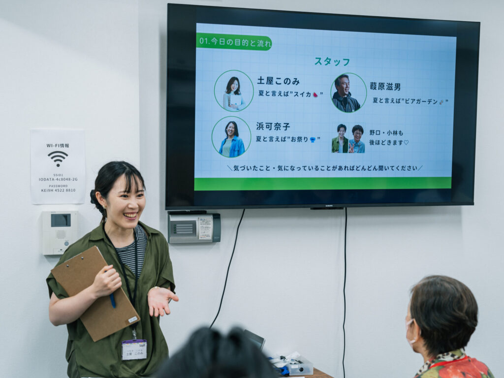 研修室前方のスクリーンの横に立ち、参加者に向かって話す土屋さん。スクリーンには、研修スライド「スタッフ紹介」が映し出されている。