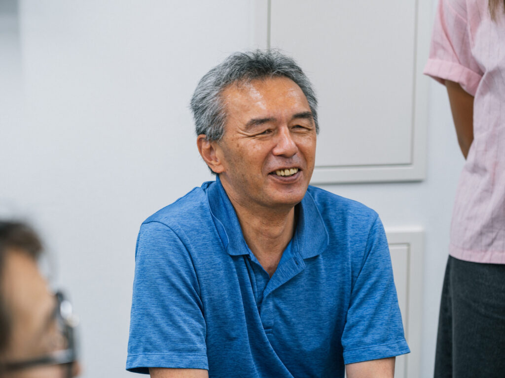 壁を背にして座っている、笑顔の葭原さん。