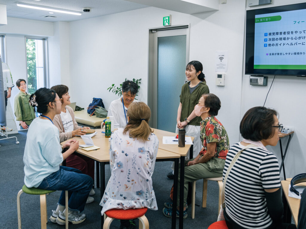 机を真ん中に、輪になってイスに腰掛ける5人の参加者。そばに立つスタッフと笑って会話している。
