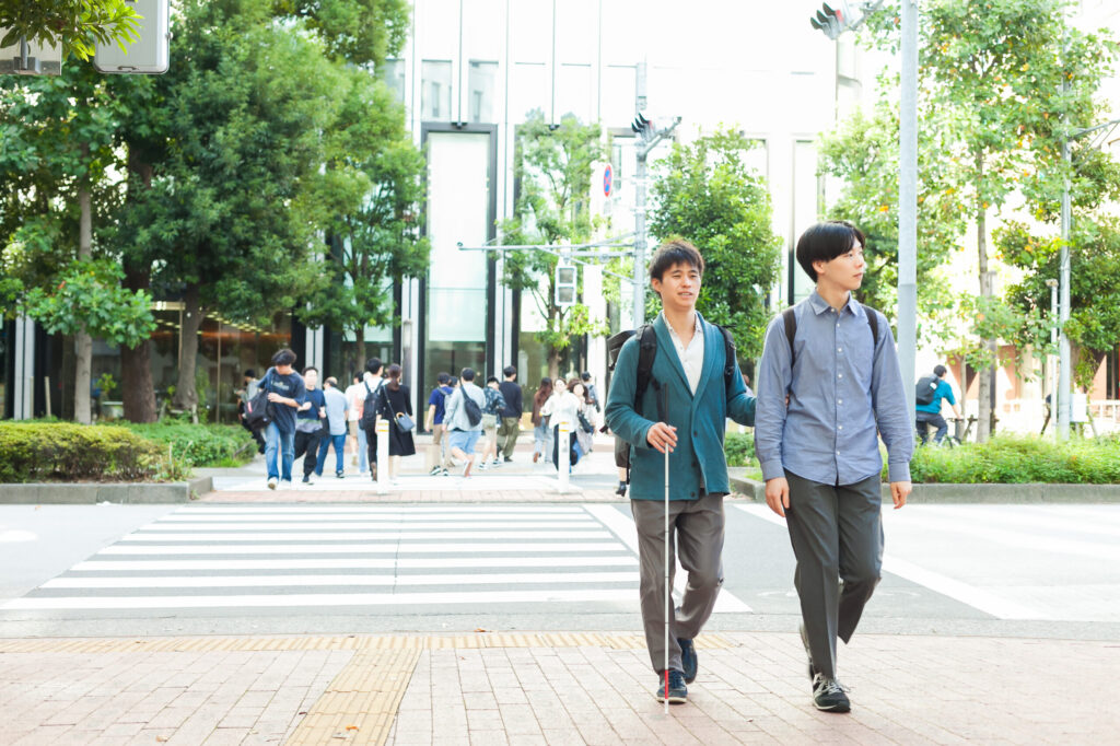 白杖の人とガイドヘルパーが、大都市の広い道路の横断歩道を渡り終えたところ。