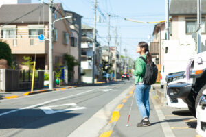 晴れた昼間の住宅街、白杖を持った女性が歩道に立って車道の方を向いている。顔をあげ斜め上を見上げている。