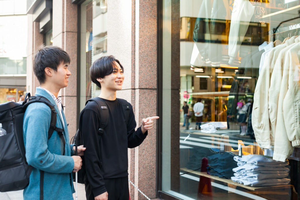 白杖の男性とガイドヘルパーの男性が、歩道から店舗のウィンドウを指さしながら会話している。ウィンドウには白いジャケットや畳まれた紺や水色のスウェットなどが見える。