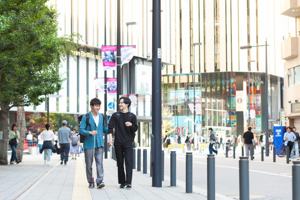大都市の繁華街の中を会話しながら歩く白杖の人とガイドヘルパー。広い歩道で、背景には百貨店など大きなビルが見える。