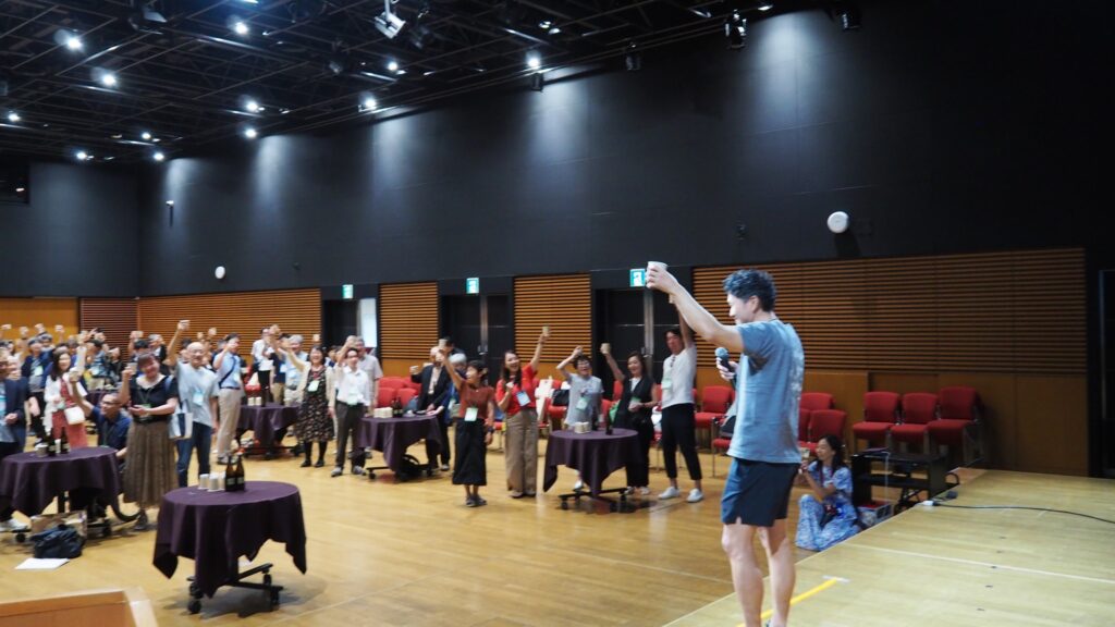 壇上で乾杯をしている鈴木直也さんを左斜め後方から写した写真。会場全体には円卓が置かれ、多くの人が飲み物を片手に笑顔で乾杯している。