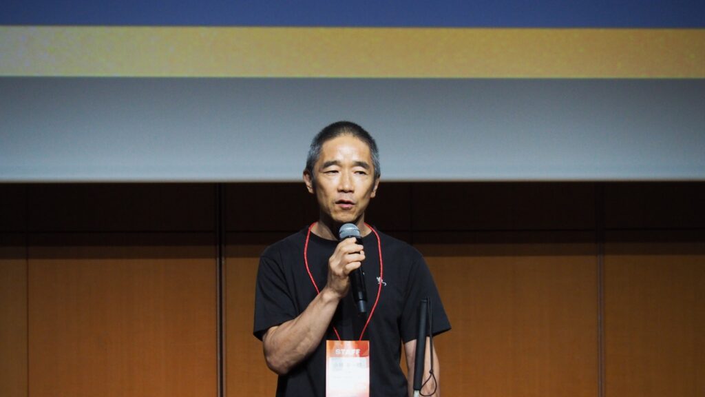 スクリーンを背に、マイクを右手に持ち話す小林さん。左手に白杖を持っている。