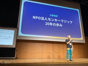 「NPO法人モンキーマジック20年のあゆみ」という言葉が映ったスクリーンと、その前に立ちマイクを持っている代表の小林さん。