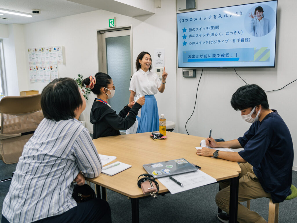 スクリーンに移された資料を指さしながら笑顔で話している野村さん。白いブラウスに明るい水色のプリーツロングスカートを着ている。手前の円卓には3名の参加者が見える。