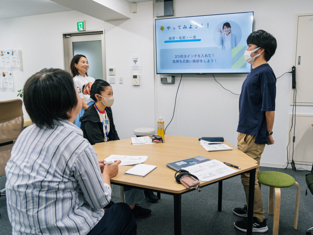 研修参加者のヘルパーの一人が立ち上がって挨拶のロールプレイをしている。講師の野村さんは笑顔で聞き、他の参加者は座って真剣に聞いている。