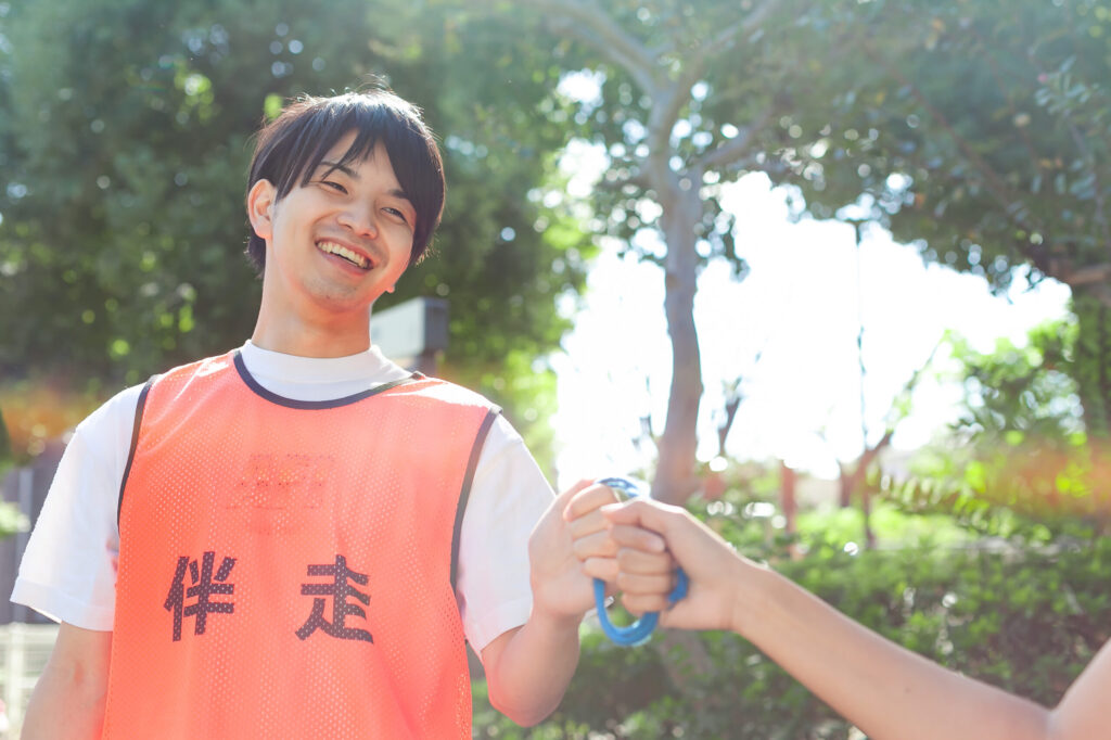 伴走と書かれたオレンジ色のビブスを着たガイドランナーが輪になったロープを左手に持ち、視覚障害者の右手がそのロープを一緒に握っている。