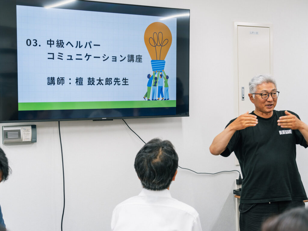黒いTシャツを着た檀鼓太郎さん。両手を胸の前に持ってきて話している。壁のスクリーンには「中級ヘルパーコミュニケーション講座/講師：檀鼓太郎先生」の文字。
