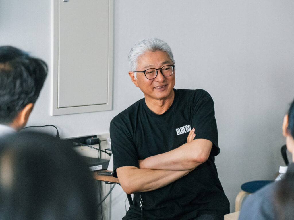 白髪で眼鏡をかけた檀鼓太郎さんが、黒いTシャツ姿で腕を組んで笑顔で話している。