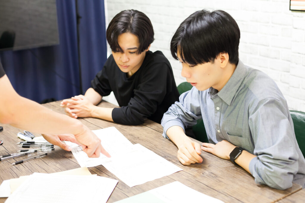 男性二人が座っていて、机に広げられた書類を見真剣な表情をしている。書類を指さしている別の人の手も見えている。