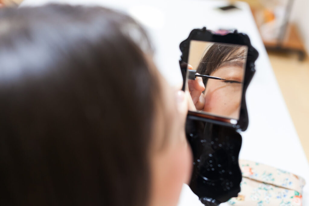 メイクしている視覚障害者女性の後姿。持っている手鏡に左目だけが写っている。丁寧にアイライナーを引いている。