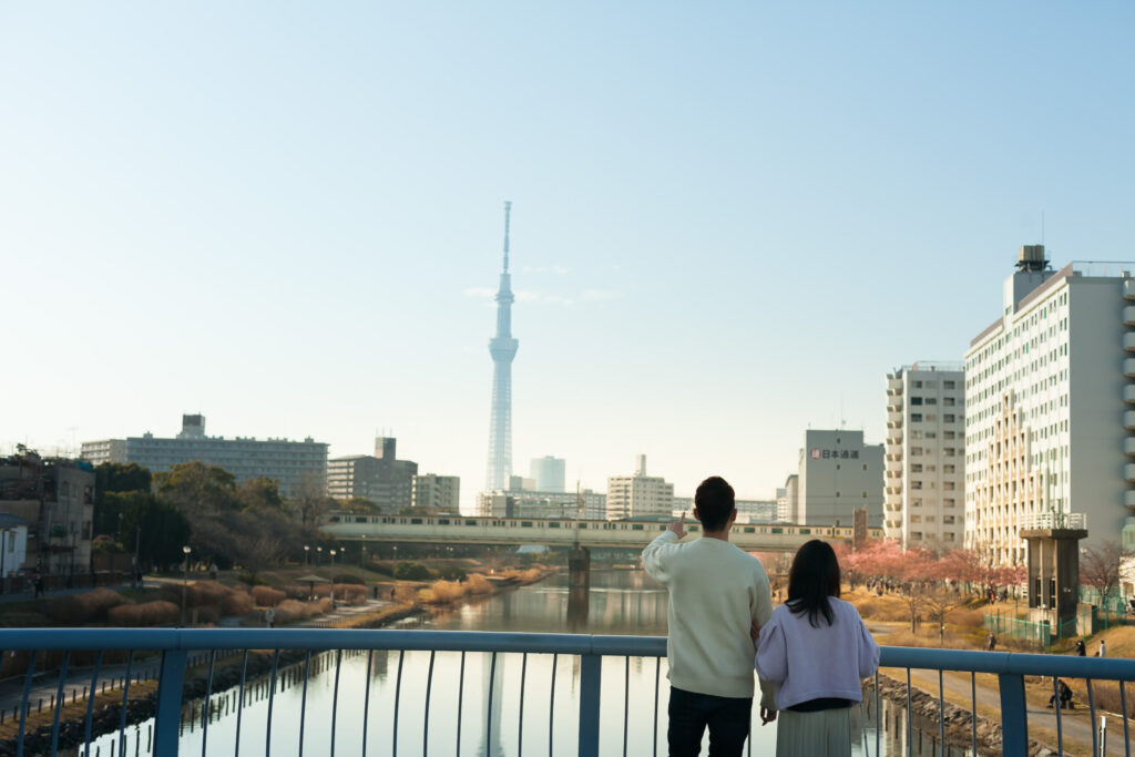 川にかかる橋の上で、遠くに見えるスカイツリーを指さしている高橋さんの後姿。視覚障害者の女性が一緒に立っている。