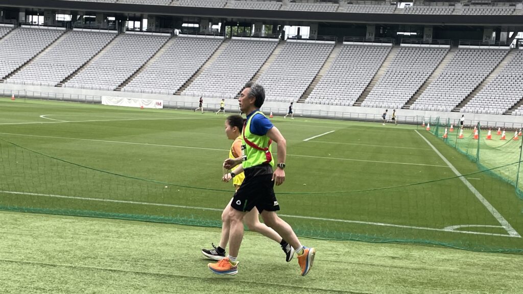 視覚障害者の男性ランナーと女性のガイドランナーが、青い芝の競技場のトラックを一緒に走っている。